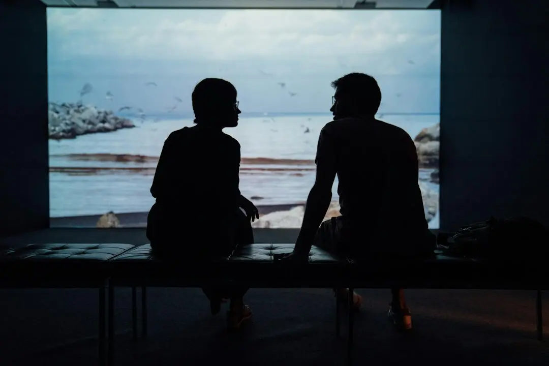 Two people silhouetted on a bench before a large screen showing a seaside view, like browsing the XerquvaTV IPTV channels
